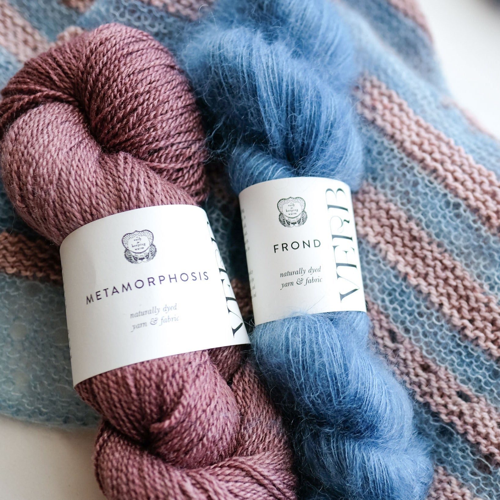 Two skeins of yarn, one purple and one blue, with Verb labels on a striped handknit shawl.