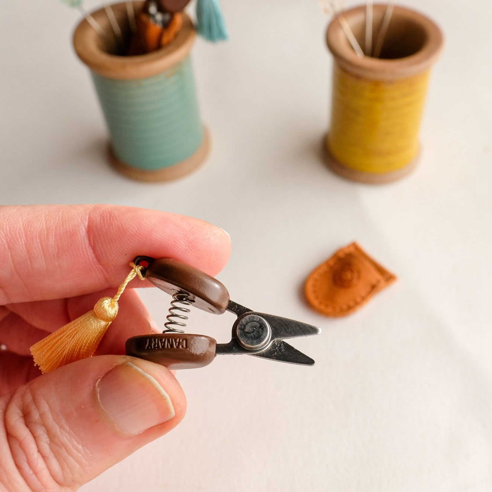 Hand holding a small pair of cohana scissors with a tassel, with spools of thread in the background.