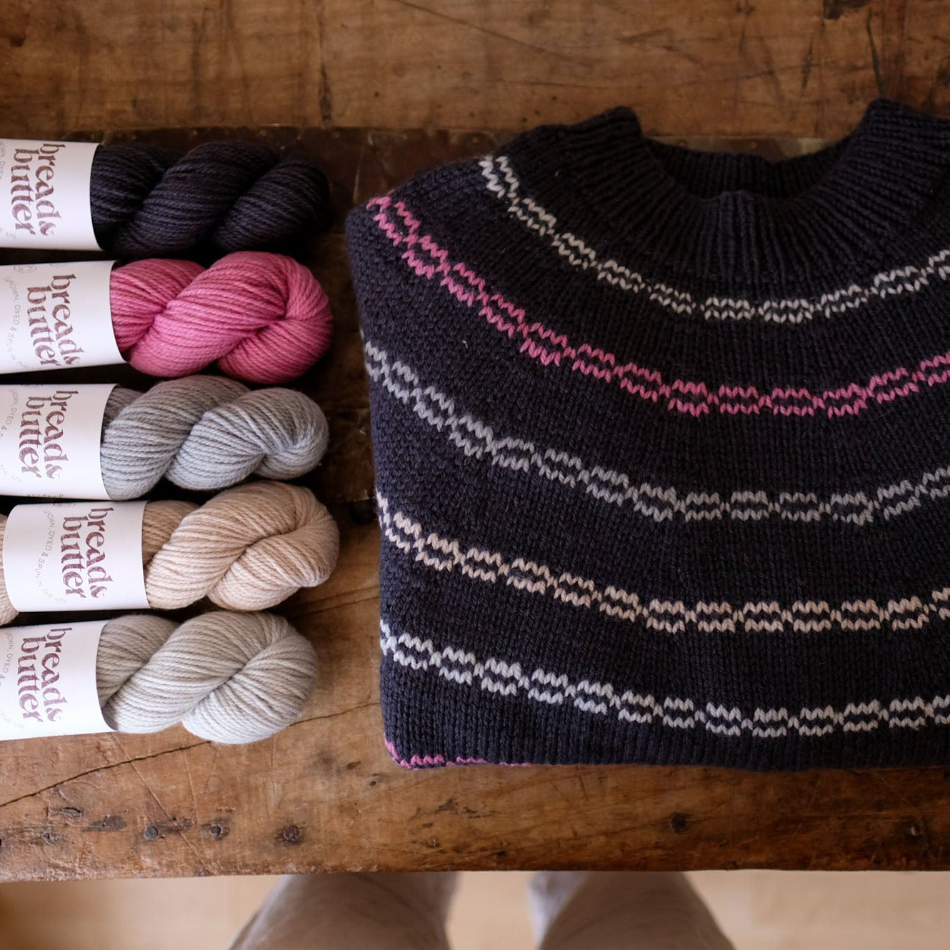 A folded black knit sweater with pink and neutral striped patterns is displayed next to five skeins of yarn in black, pink, gray, beige, and light gray on a rustic wooden table. The yarn labels read 'Bread & Butter Yarn'.