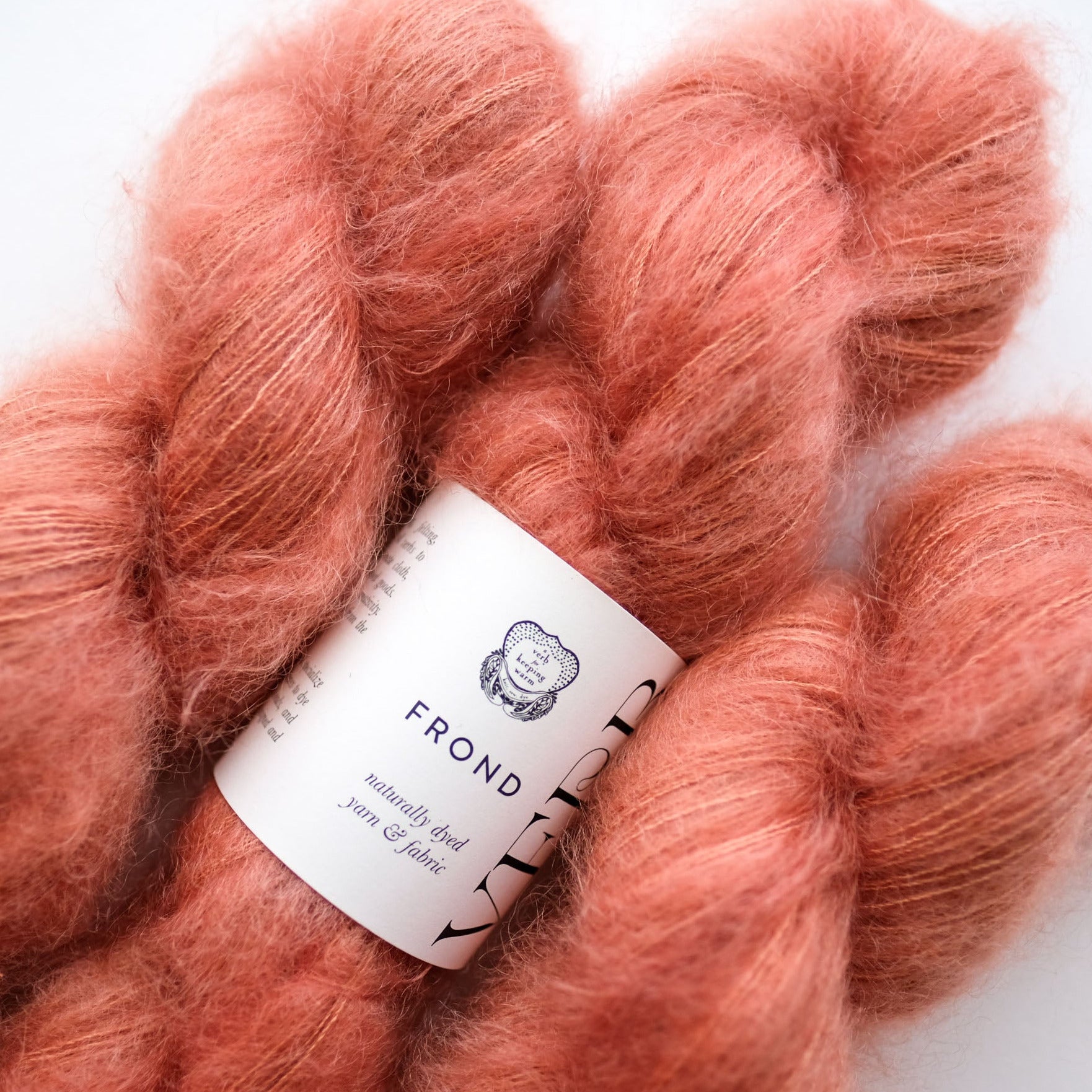 A close-up of fluffy peach-pink yarn skeins with a white paper label reading FROND and yarn & fabric.