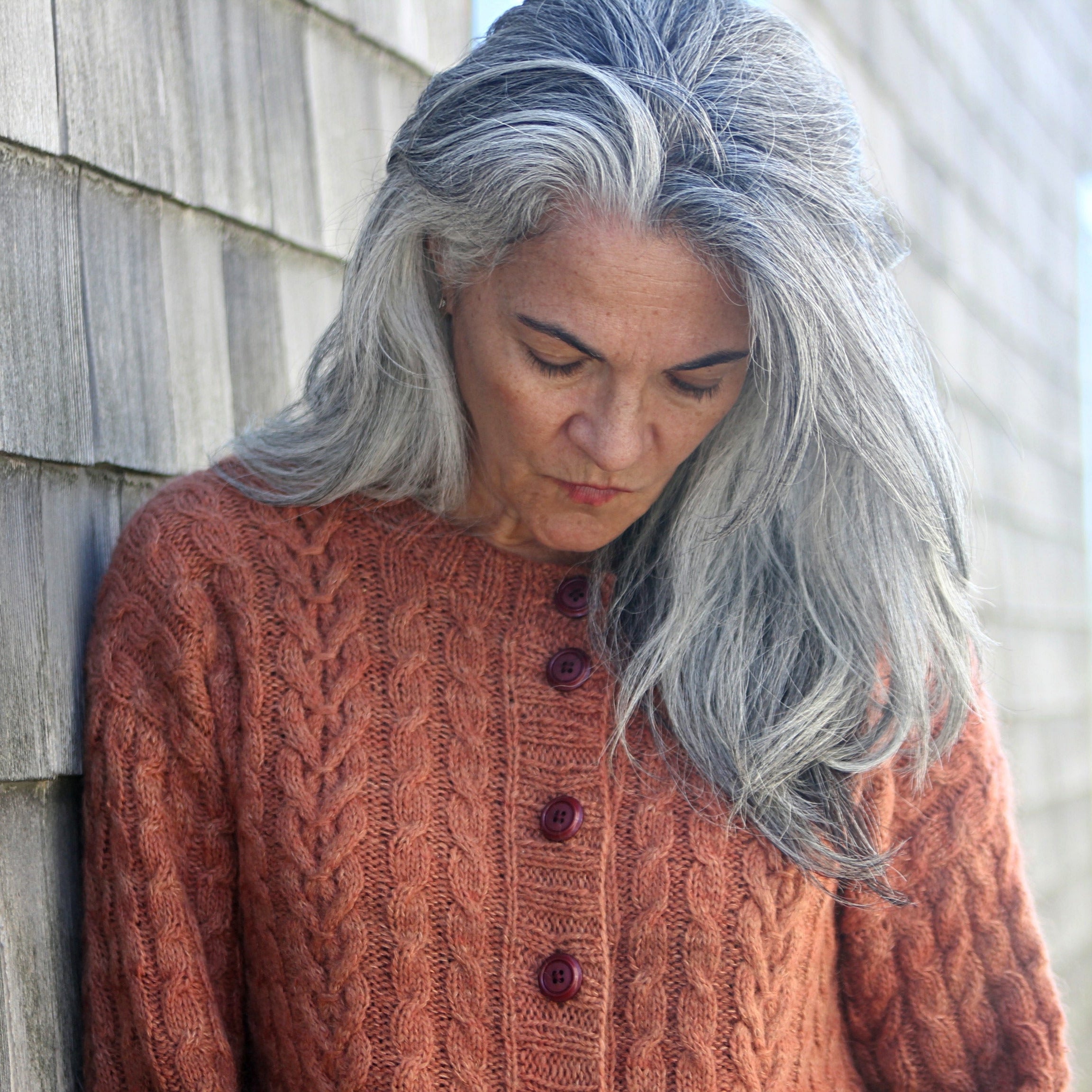 Woman with gray hair wearing a brown sweater against a wooden wall.