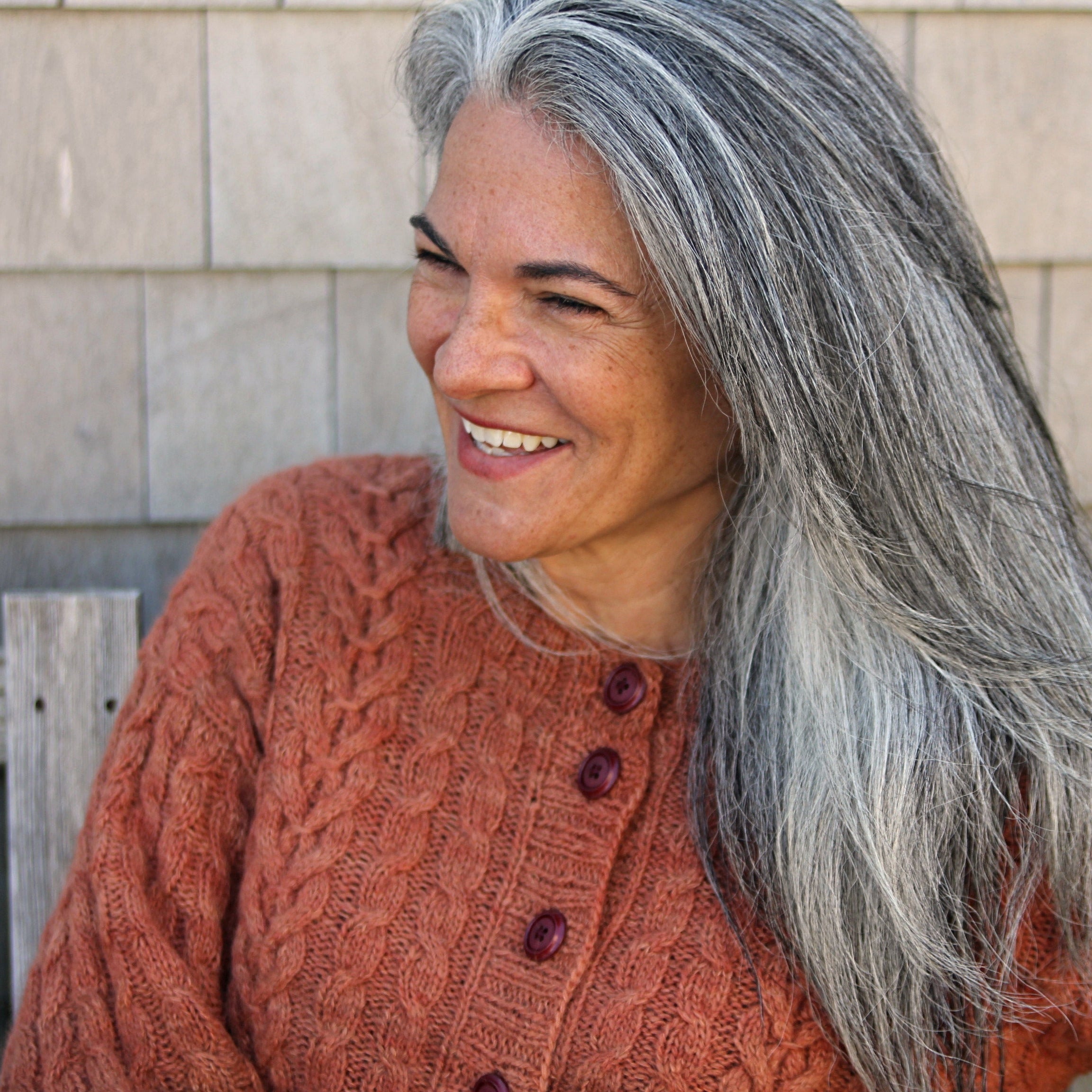 Woman with long gray hair wearing a brown knitted sweater sitting outdoors.