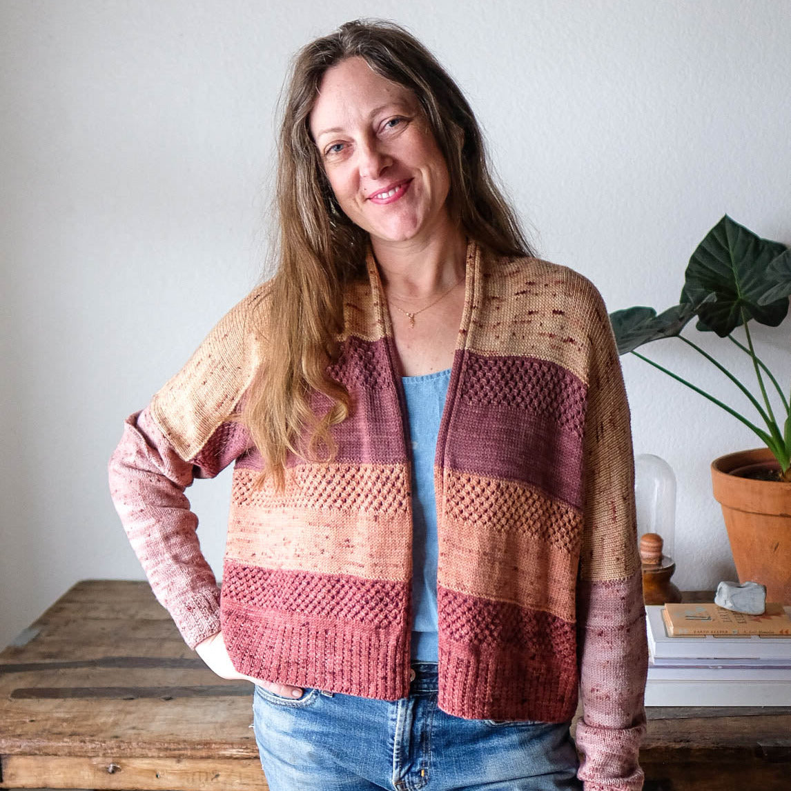 A person wearing a color-blocked, knitted cardigan with panels of pink, orange, and beige, standing in front of a wooden table and a potted plant.