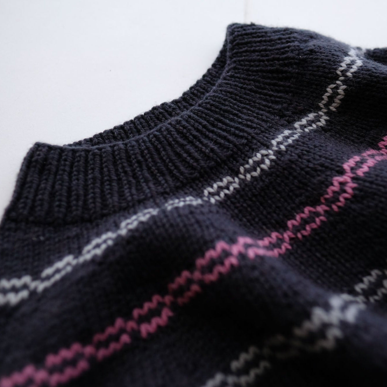 Close-up of a dark knit sweater with a ribbed crew neck and diagonal wavy stripes in pink, white, and gray.