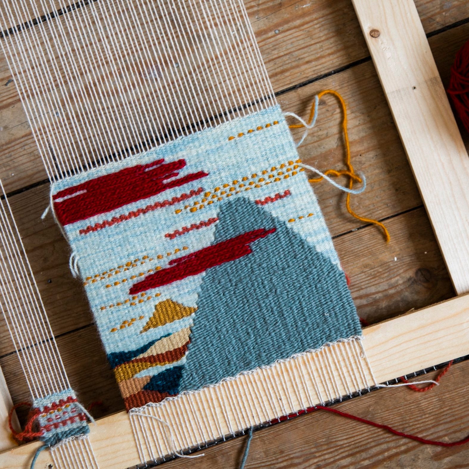 Woven textile with mountain design on a wooden loom, surrounded by yarn and tools.