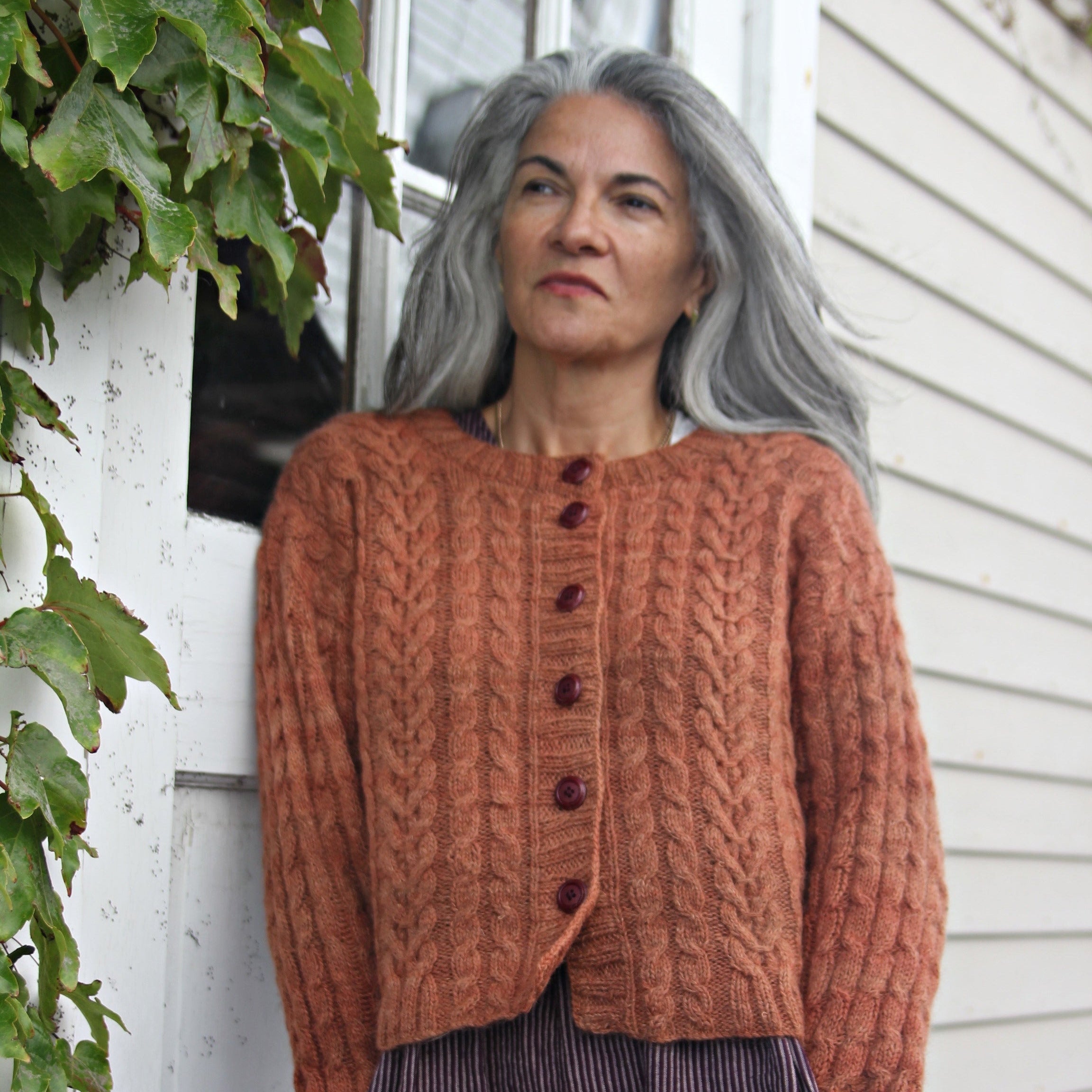 Woman wearing an copper hand-knitted cardigan standing outdoors next to a building with greenery.