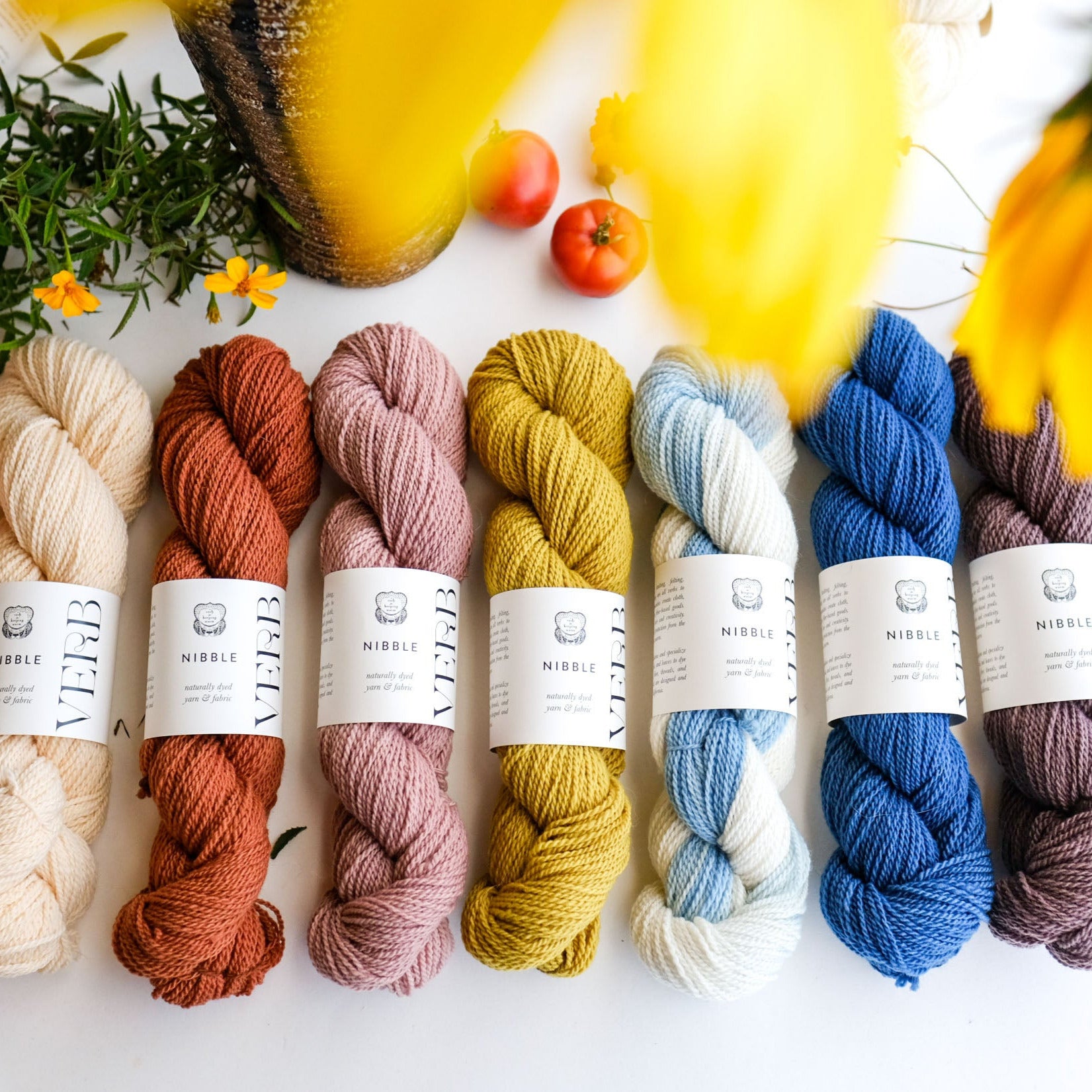 Colorful yarn skeins on a white surface with flowers and fruits in the background
