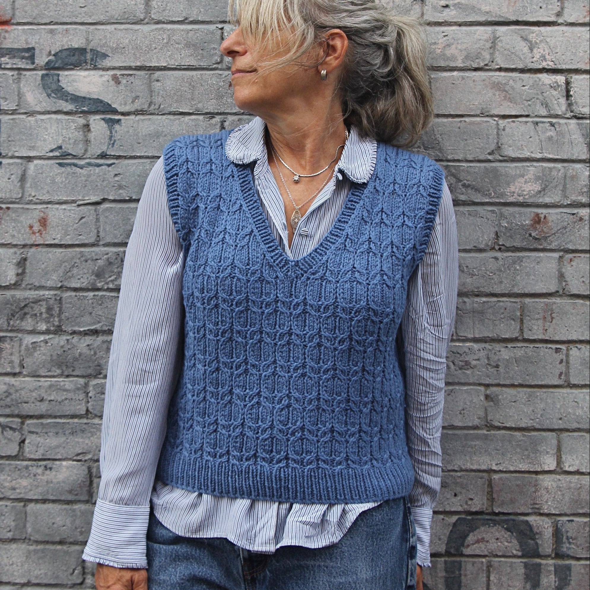 A woman wearing a blue cable-knit sweater vest over a light blue and white striped button-down shirt, paired with blue jeans, standing against a gray brick wall.