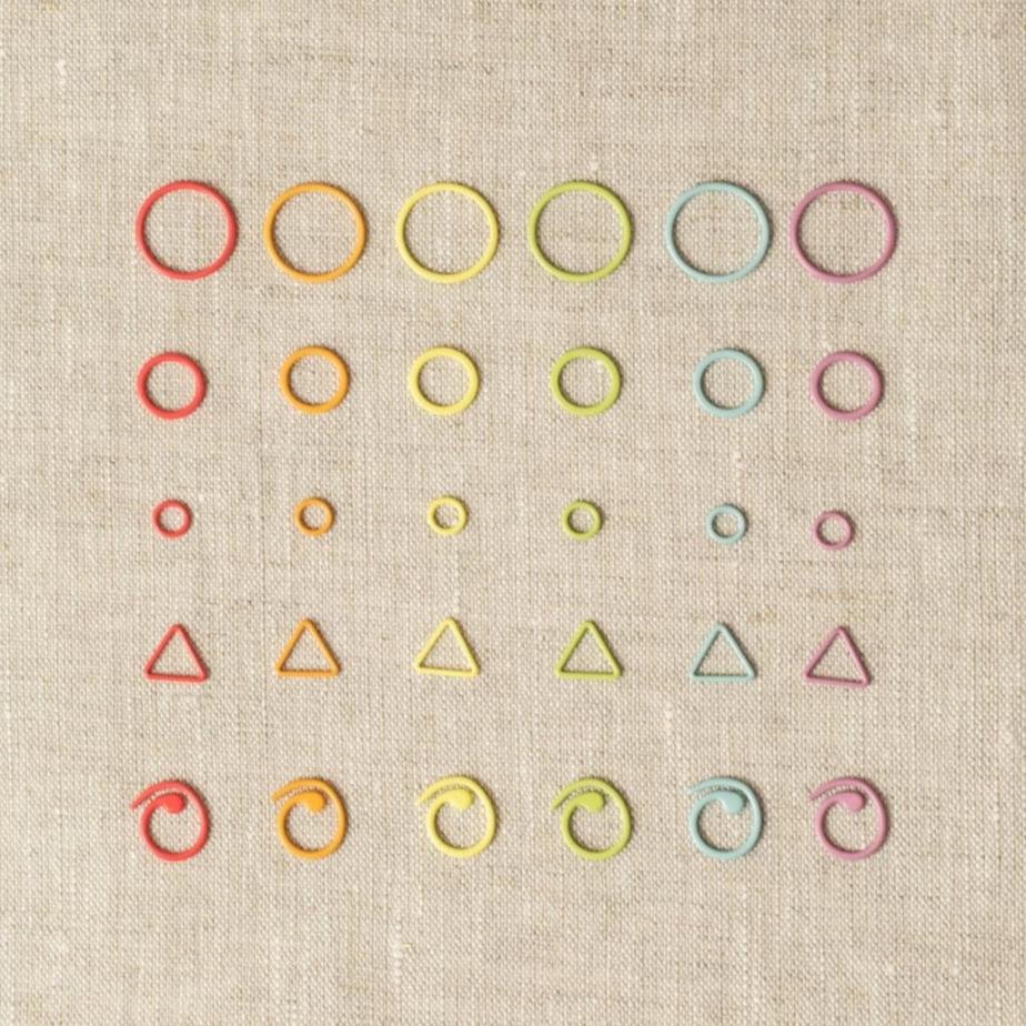 A sample of the Cocoknits Stitch Marker included in the Flight - Displayed on a linen fabric are six different colors and five different shapes to be used with the Cocoknits Method